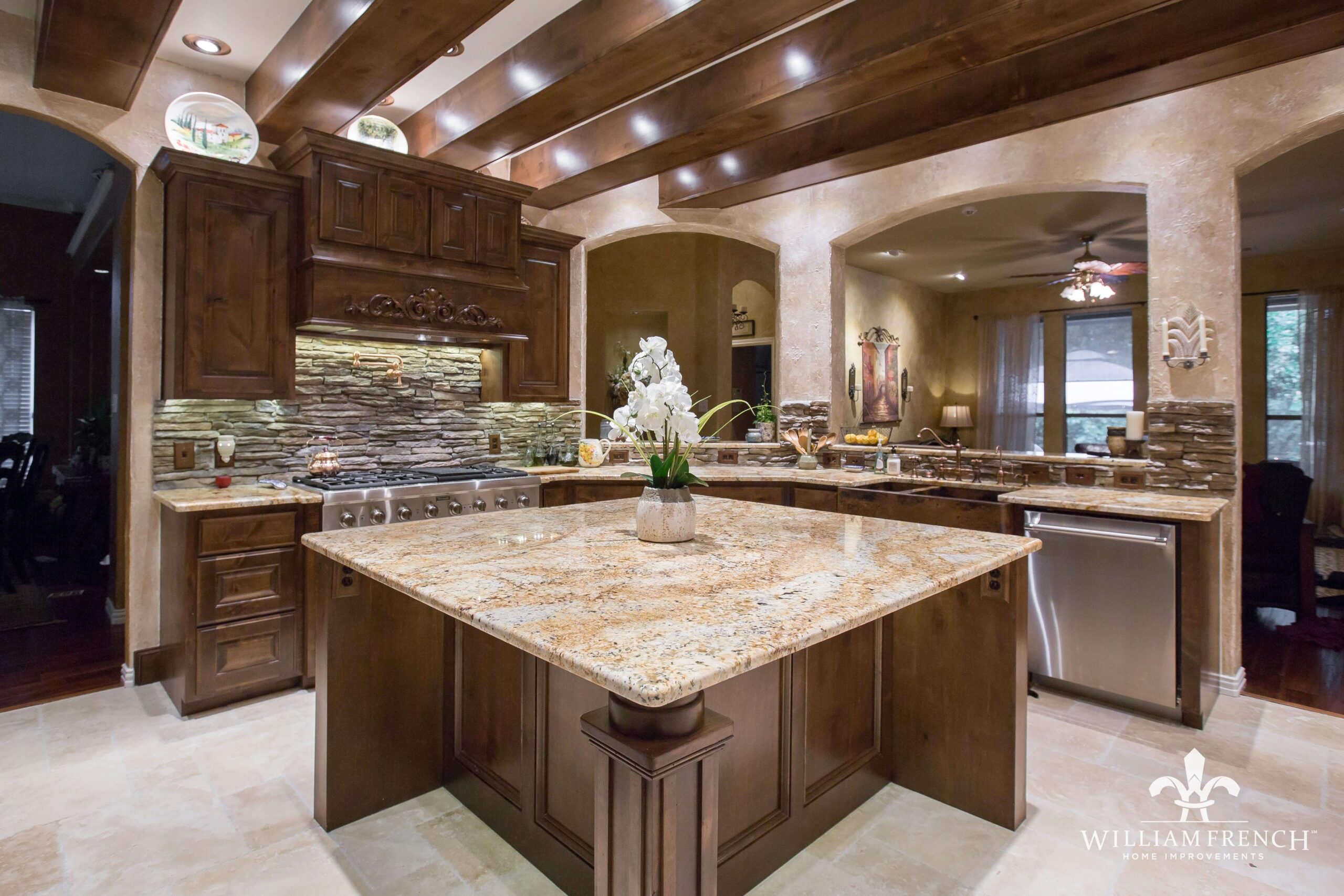 A spacious, elegant kitchen featuring wooden cabinets, a large central island with a granite countertop, and a modern stainless steel stove. The back wall displays a stone backsplash, and a white orchid decorates the island. The ceiling has wooden beams.