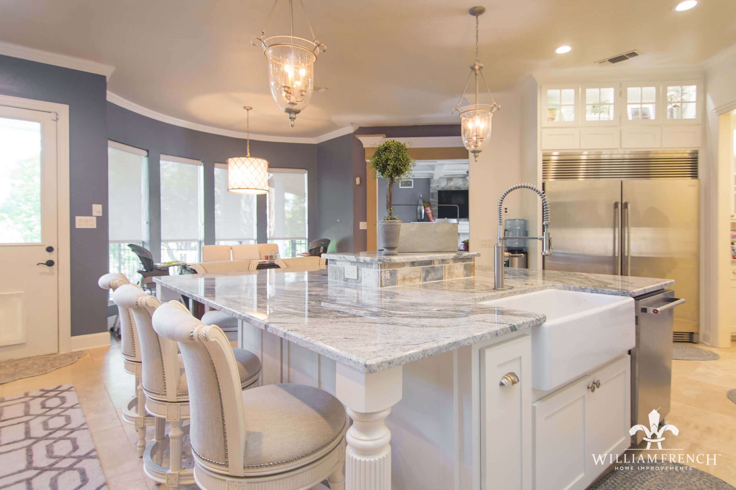 A modern kitchen features a large island with a marble countertop and cushioned stools. Stainless steel appliances, including a refrigerator and oven, are visible. Elegant pendant lights hang above the island, and large windows offer ample natural light.