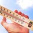 A hand holds a wooden thermometer against a bright blue sky, showing high temperatures in both Celsius and Fahrenheit—a reminder of the importance of choosing the best insulation for hot climates.