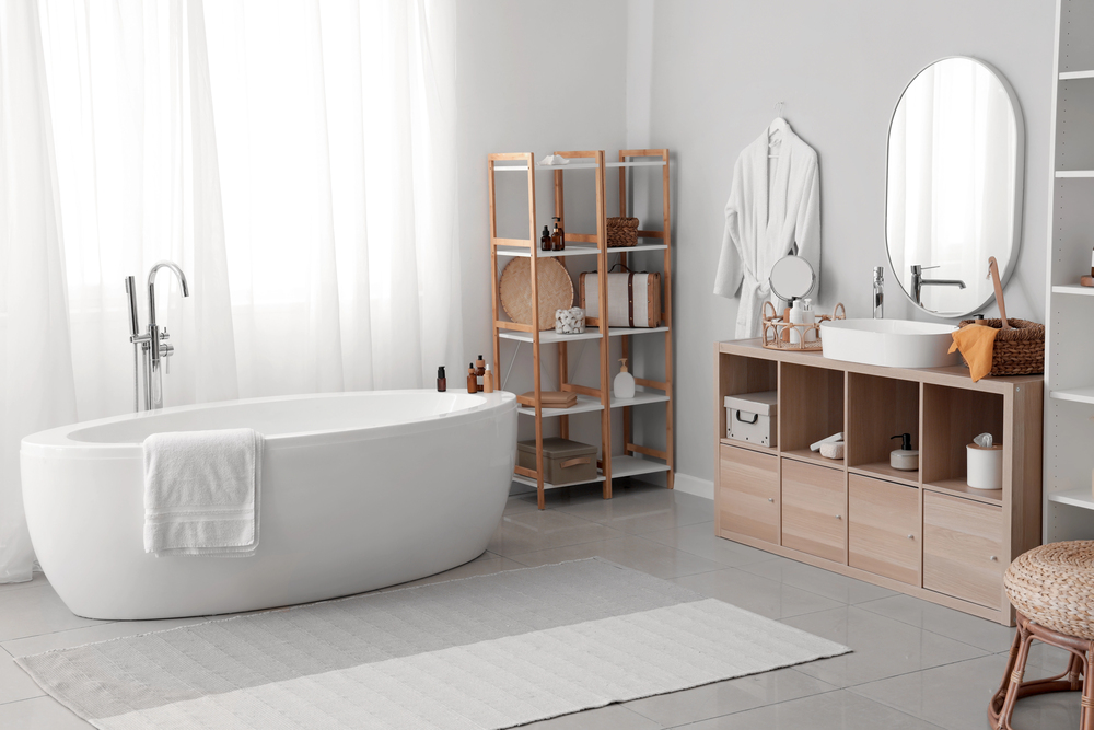 Modern bathroom with a white freestanding bathtub, a towel draped over the side, sleek bathroom tiles, wooden shelving with baskets and decor, a vanity with sink, round mirror, and a robe. Light streams in through sheer curtains.
