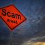An orange diamond-shaped road sign with the words "Scam Ahead" stands against a dark, cloudy sky at sunset, warning of potential contractor scams or danger from shady contractors ahead.