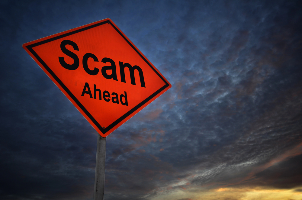 An orange diamond-shaped road sign with the words "Scam Ahead" stands against a dark, cloudy sky at sunset, warning of potential contractor scams or danger from shady contractors ahead.